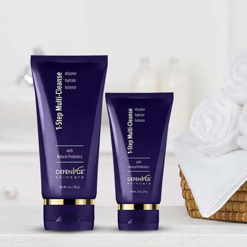DefenAge 1-Step Multi Cleanse full and travel size tubes on vanity beside towels for hydrating daily face cleansing routine.