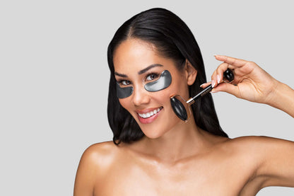 Model wearing black Knesko eye masks while using facial roller for a smoother refreshed under eye skincare look at home use.