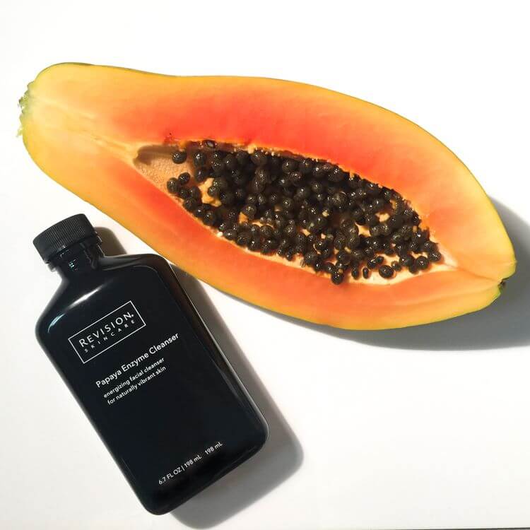 Revision Skincare Papaya Enzyme Cleanser bottle placed beside fresh papaya fruit highlight enzyme based exfoliating formula