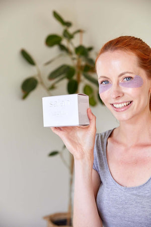Saian ActiveCell Collagen Stem Cell Eye Pads model holds box while wearing purple under-eye patches for spa care.