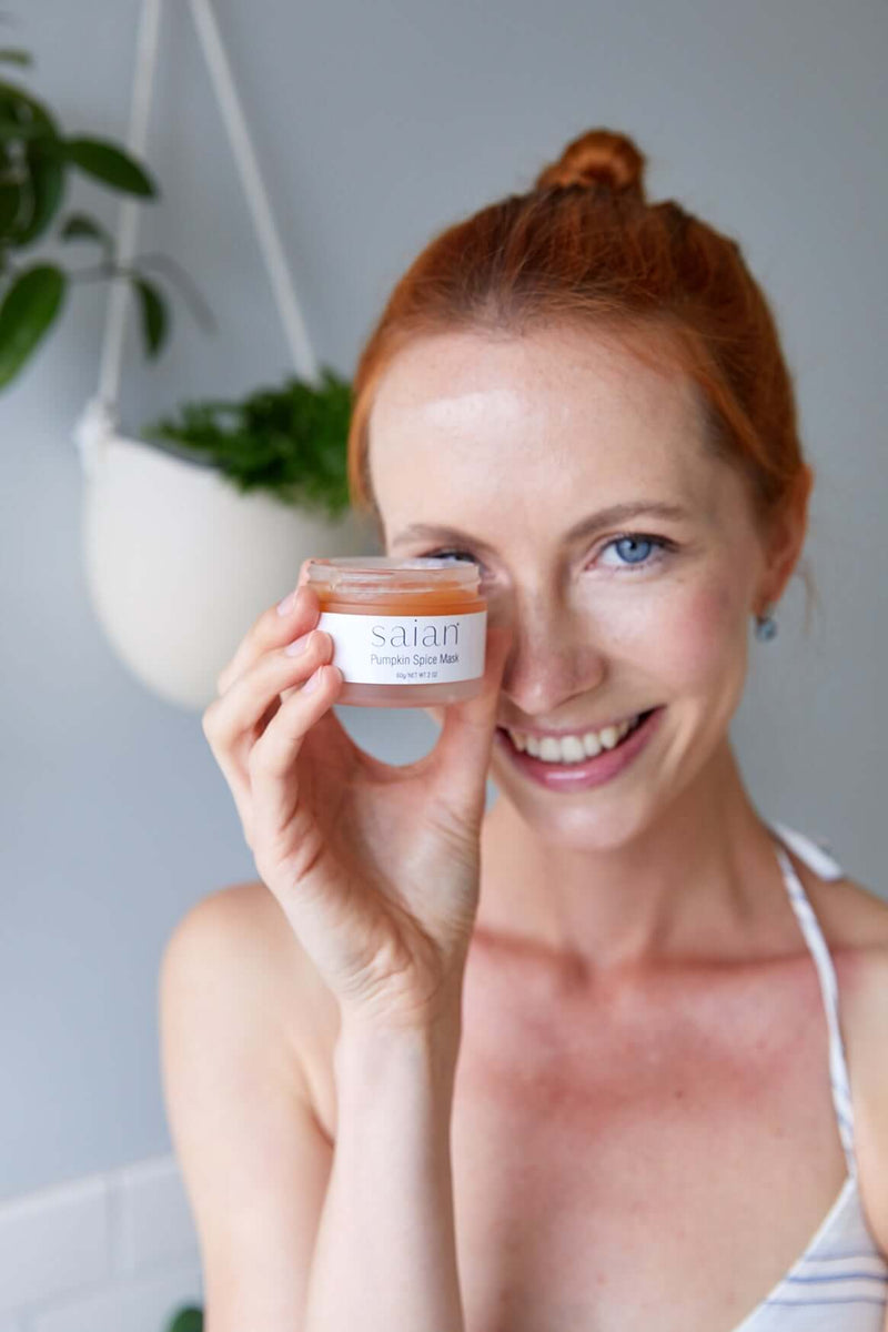 Saian Pumpkin Spice Mask held near face by model for brightening, smoothing skincare mask product lifestyle image.